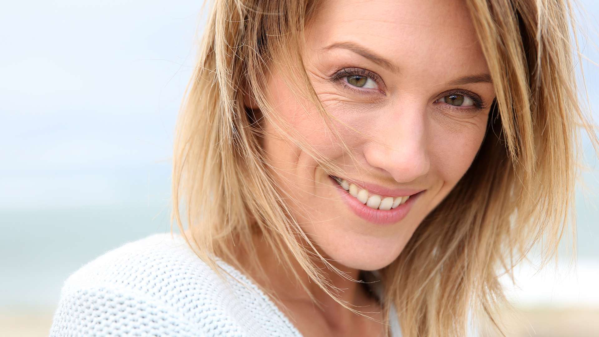 A woman with shoulder-length blonde hair and light eyes smiles warmly at the camera, wearing a white sweater. Her inviting expression hints at confidence—perfect for learning How To Get An Older Woman In The Mood in a natural, outdoor setting.